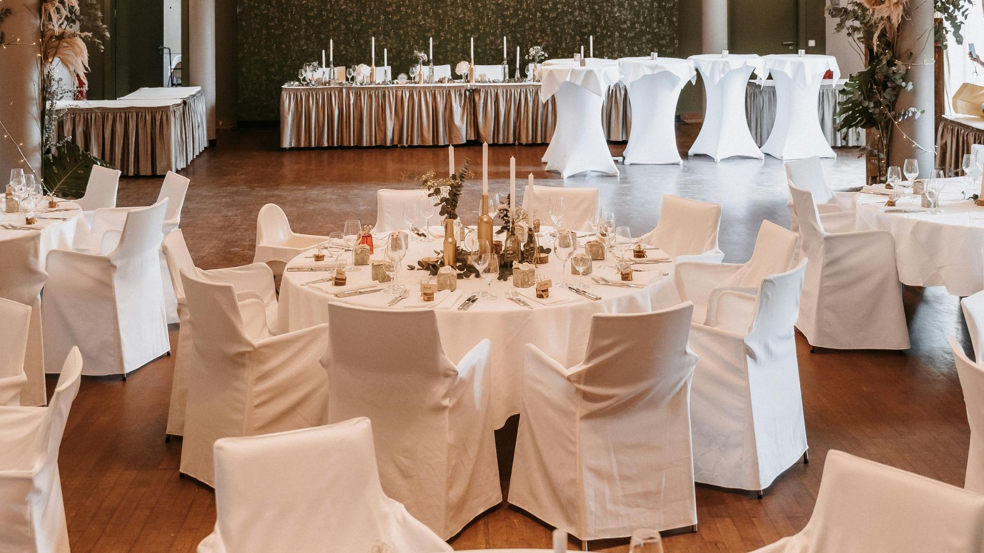 Elegant banquet hall prepared for a formal event, featuring large round tables covered in white linens and surrounded by chairs with white slipcovers